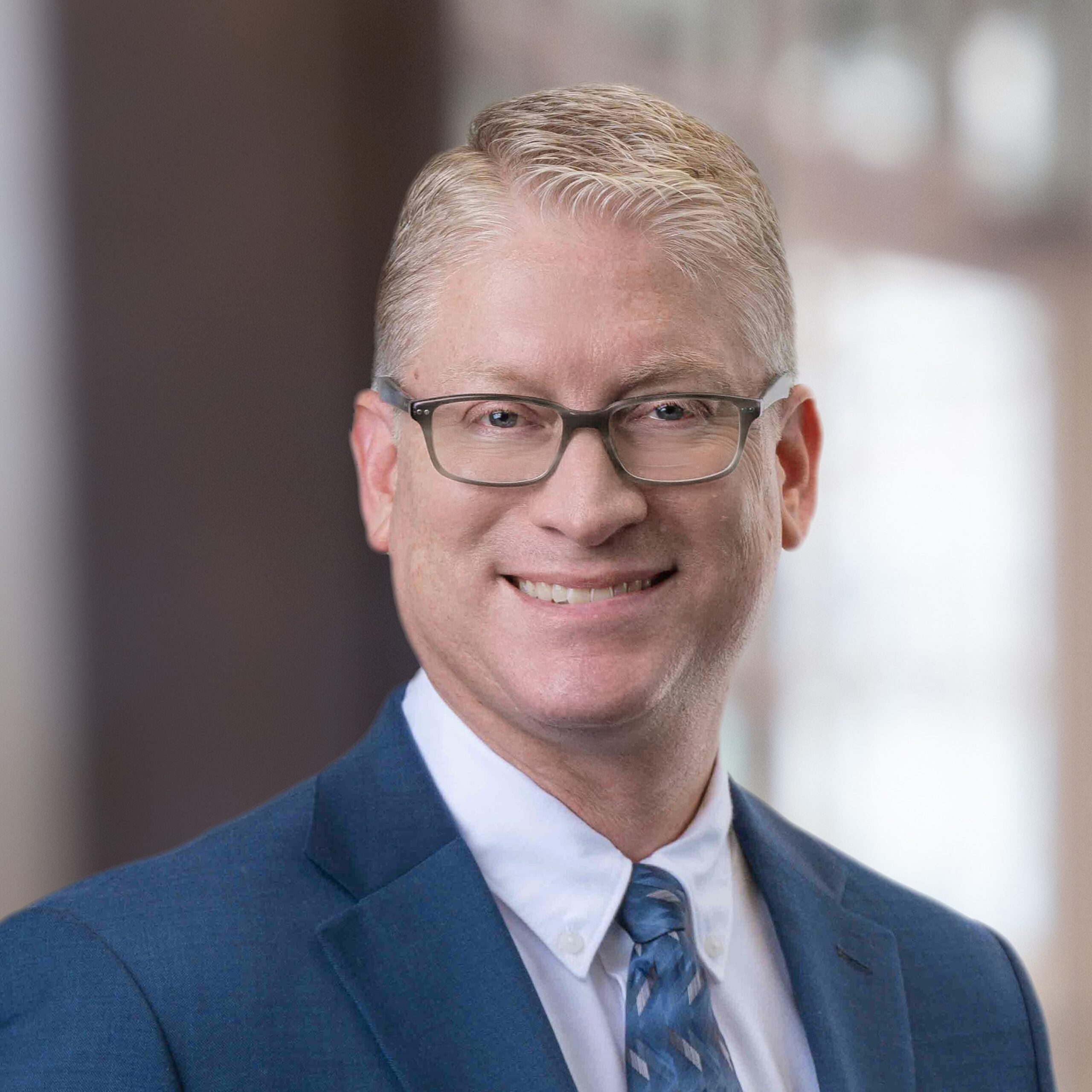 Headshot of a middle aged blond man in glasses, suit jacket, and tie smiling in front of a blurred indoor background