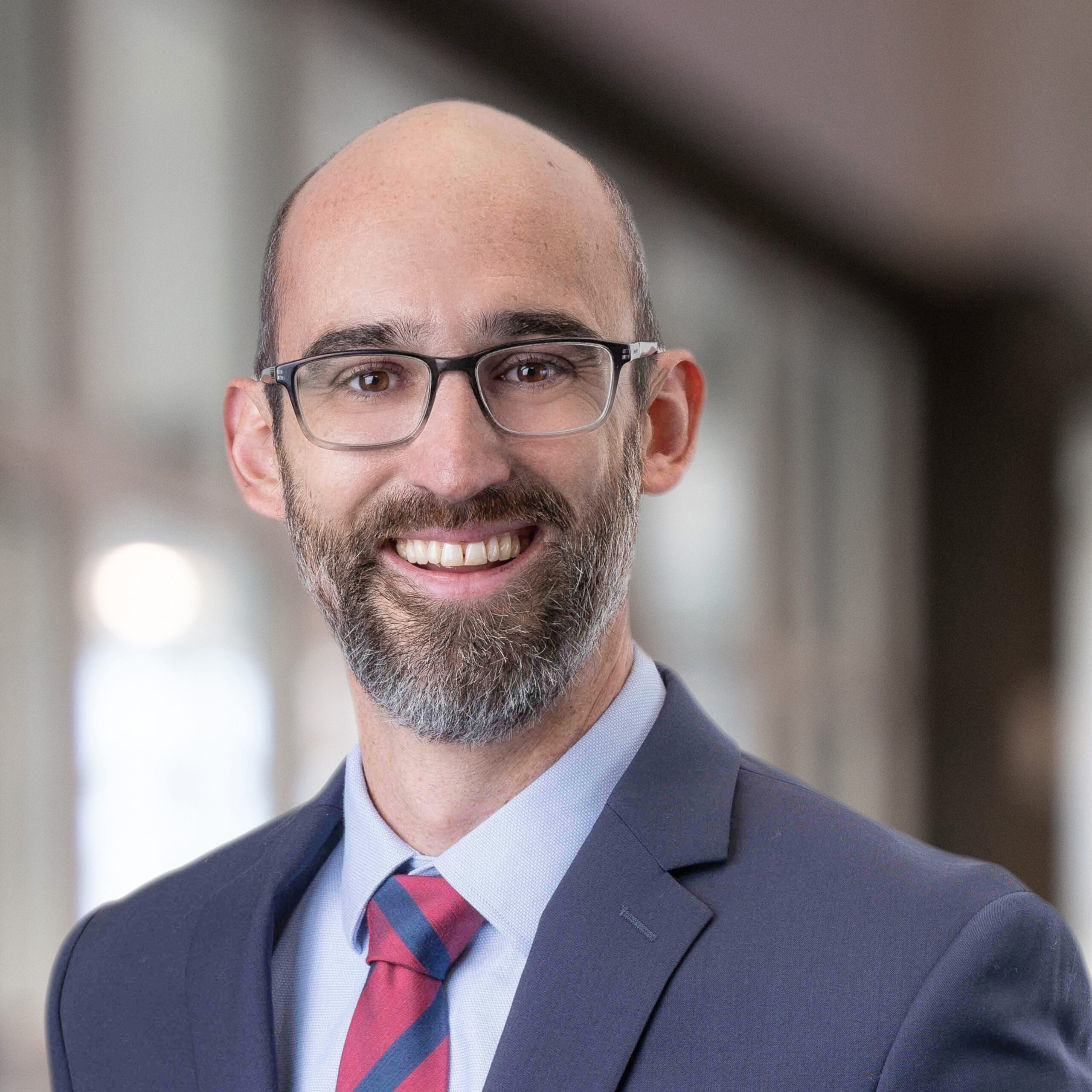Headshot of a bearded middle aged man with glasses, suit jacket, and tie smiling in front of a blurred indoor background