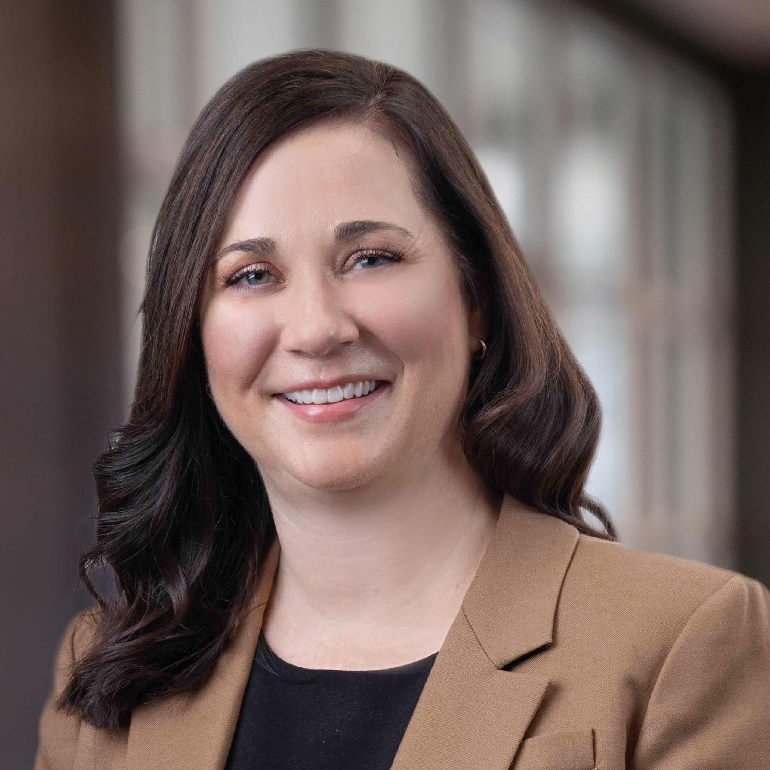 Headshot of a dark haired woman in a brown blazer smiling for the camera in front of a blurred indoor background